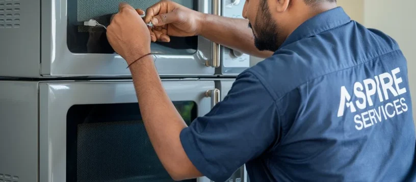 An Aspire technician in a blue uniform carefully inspecting a grey microwave oven for quality and performance.