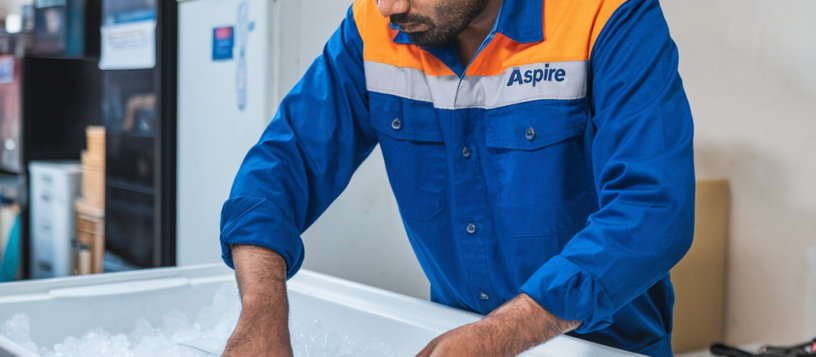 Aspire technician in a blue uniform working on a white freezer, focusing on repairs with precision and expertise.