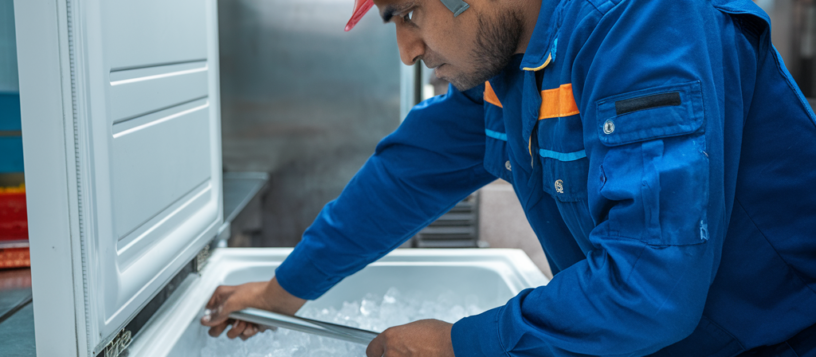 Aspire technician wearing a white helmet and blue uniform reviewing a white freezer box, focusing on assessing the appliance for repairs. Freezer Repair Service Mysore